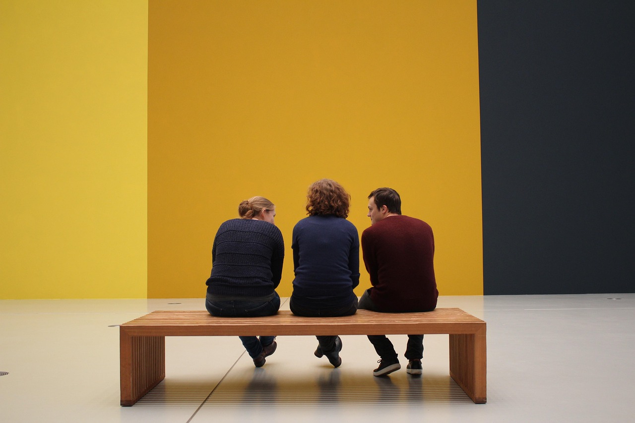 Trois personnes assises de dos sur un banc en bois dans un musée aux murs jaunes et noirs